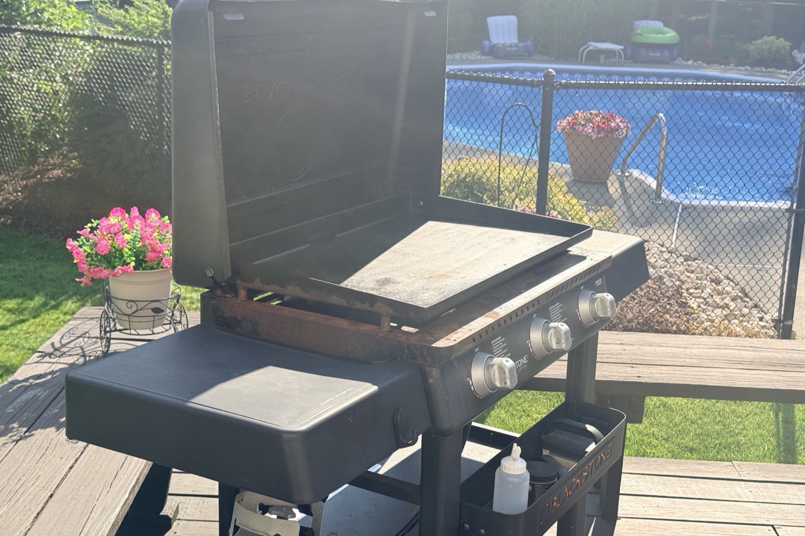A well-used Blackstone flat top griddle with the lid open and a seasoned steel plate, on a wooden pool deck