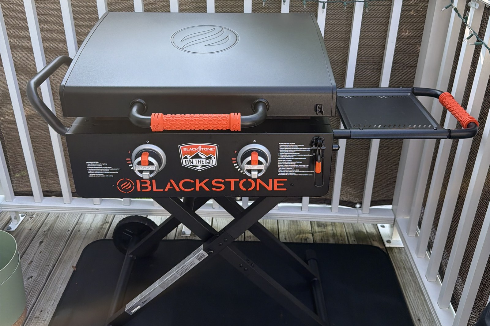A two-burner Blackstone On The Go griddle with the hood closed, folded side shelf raised, standing on scissor legs with wheels on a wooden deck